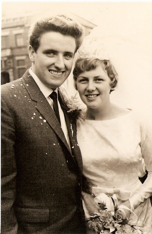 Sepia-toned image of a young, newly-married couple on their wedding day. On the left is a man with dark hair in a quiff, and wearing a dak suit with a white shirt and black tie.On the right is a slightly shorter woman, with light hair covered in a thrown-back veil, and wearing a white, heavily embroidered wedding dress, holding a posey of flowers at waist level. Both are looking at the camera and smiling.
